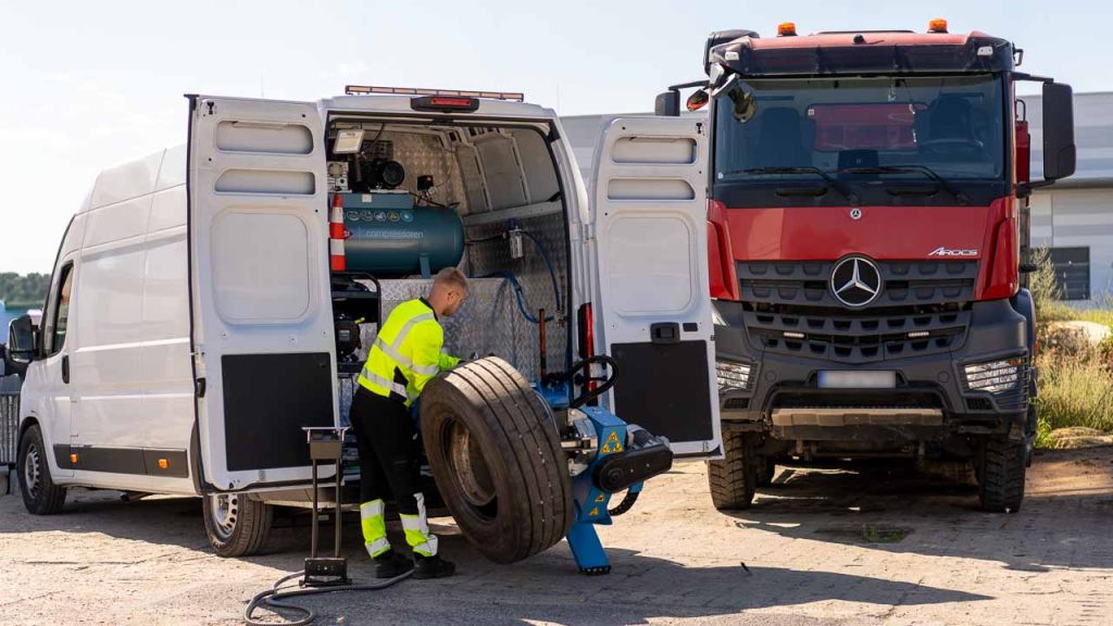 Serwisant mobilnej wulkanizacji obsługuje koło bliźniacze na montażownicy w busie, obok czerwona ciężarówka Mercedes Arocs