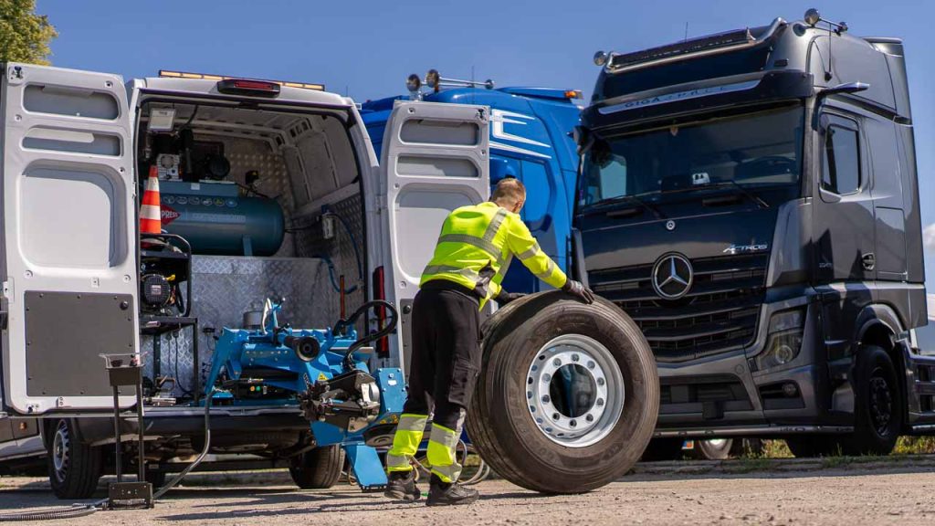 Serwisant przetacza koło tira z nową oponą do szarego Mercedesa Actros, obok bus serwisowy z montażownicą